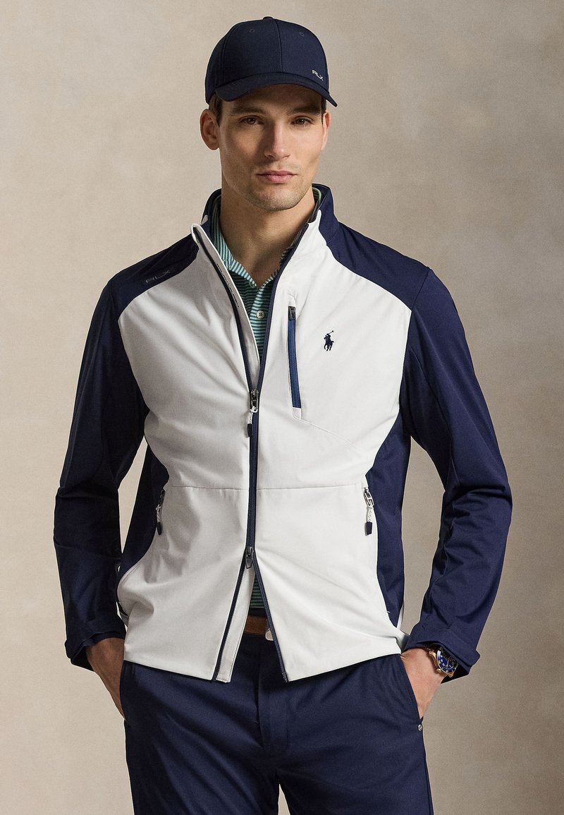 Muž nosící námořnickou čepici, bílou a námořnicky modrou zipovou bundu přes zelené pruhované polo, s námořnicky modrými kalhotami a rukama v kapsách.
