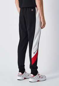 Black sweatpants with white and red side panels, featuring an elastic waistband and a logo patch on the left hip. Smooth fabric texture.