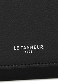 Pelle nera tridimensionale con testo in rilievo "LE TANNEUR 1898" in bianco. Presenta cuciture visibili lungo i bordi e una finitura liscia.
