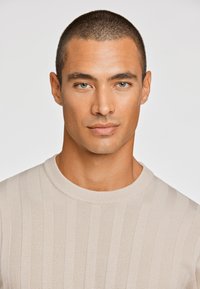 Ανοιχτό μπεζ πλεκτό πουλόβερ με ραβδωτή υφή και στρογγυλή λαιμόκοψη. Το ύφασμα έχει ένα διακριτικό σχέδιο με εναλλασσόμενες ρίγες.