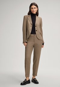Costume à chevrons marron avec une veste ajustée et un pantalon fuselé, associé à un col roulé noir à côtes et des mocassins noirs avec des éléments en or.