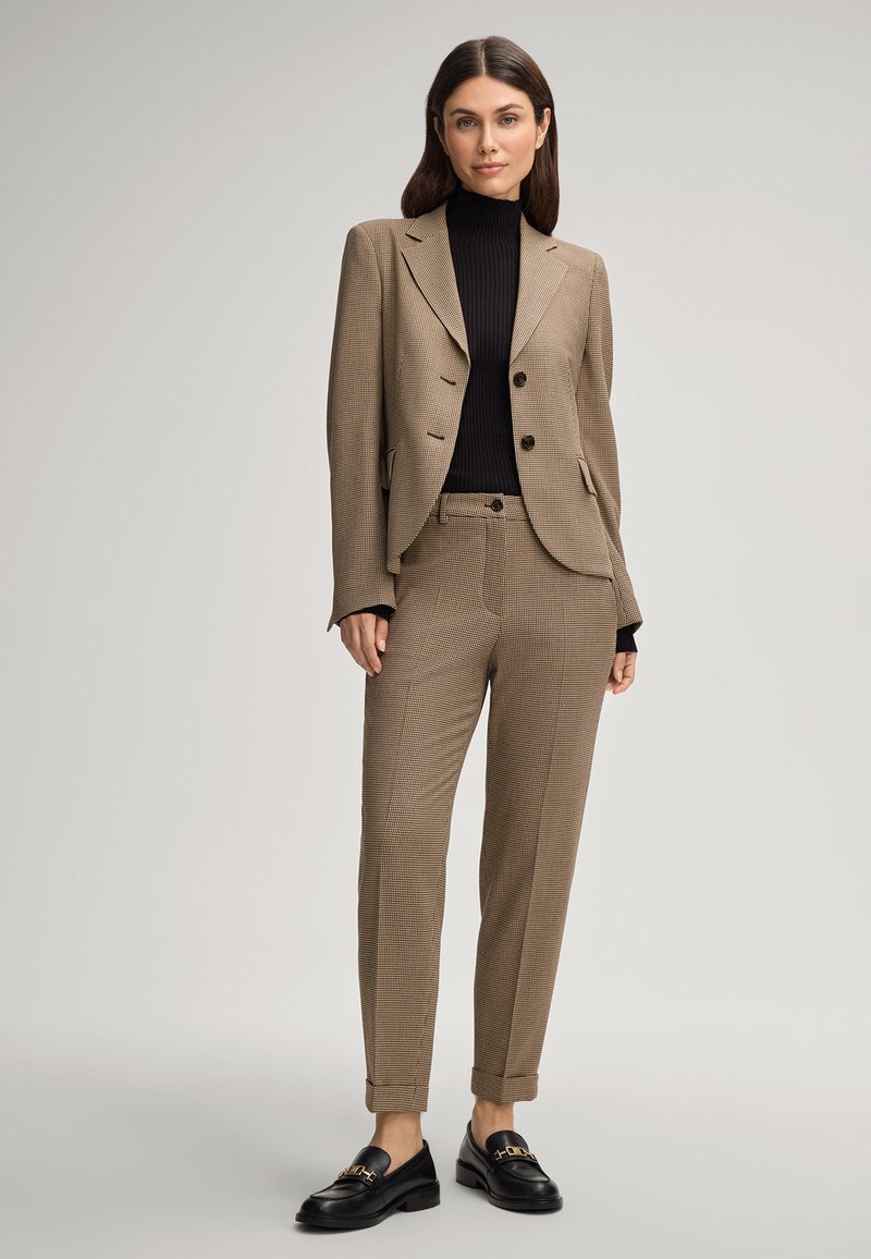 Costume à chevrons marron avec une veste ajustée et un pantalon fuselé, associé à un col roulé noir à côtes et des mocassins noirs avec des éléments en or.