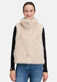Gilet en fausse fourrure de couleur beige clair avec un col haut et une surface texturée et duveteuse; porté sur un haut à manches longues noir et associé à un jean bleu.