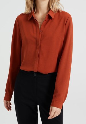 Femme portant une chemise à manches longues couleur rouille-orange boutonnée et rentrée dans un pantalon noir taille haute sur un fond clair.