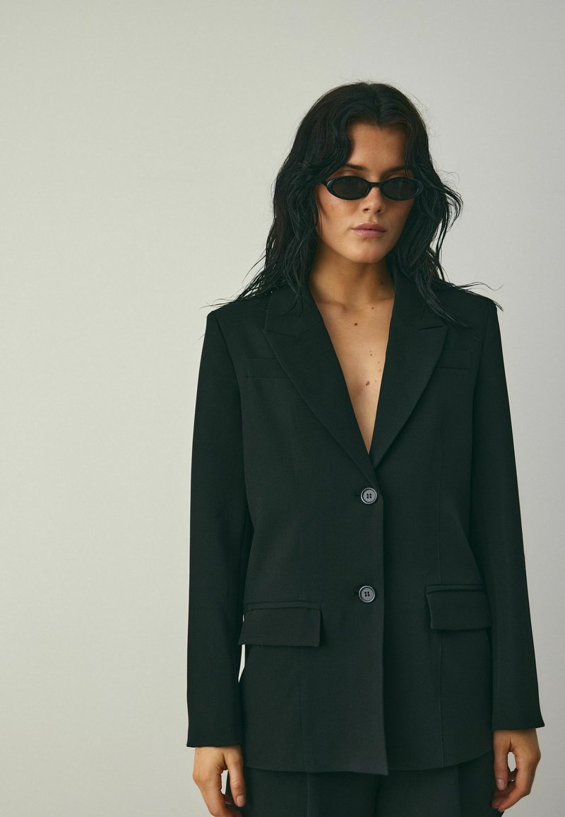 Čierny prispôsobený blejzer s dvoma prednými vreckami, klopovými goliermi a zapínaním na gombík. Hladká tkanina so štruktúrovaným siluetou.