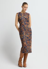 Ärmlös klänning i blått tyg med ett blommönster i bruna och gyllene toner, med en figursydd midja och fickor. Bruna knähöga stövlar.