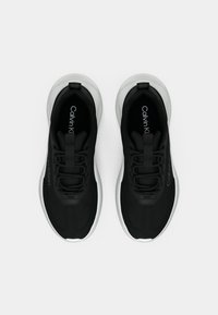 Μαύρα αθλητικά παπούτσια με γκρι σολές, υφασμάτινο επάνω μέρος και λωρίδες κορδονιών σε τονικά χρώματα. Το λογότυπο εμφανίζεται στη γλώσσα και στη πλευρά. Ομαλή υφή.