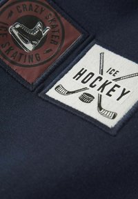 Tessuto blu navy con due toppe quadrate: una rossa con un logo di pattinaggio su ghiaccio e un'altra bianca con bastoni da hockey e un disco, entrambe ricamate.