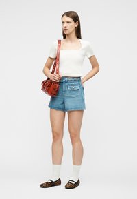 Žena oblečená v bielej krátkosleevej blúzke so štvorcovým výstrihom, modré vysoké rifľové šortky, biele ponožky, baleríny s leopardím vzorom a nesie červenú koženú kabelku na rameno.