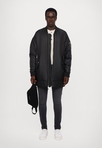 Veste bomber noire rembourrée avec des poignets en côtelé et un ourlet, portée sur une chemise henley de couleur claire. Assortie à un jean foncé et des baskets blanches.