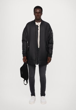 Schwarze gepolsterte Bomberjacke mit gerippten Bündchen und Saum, getragen über einem hellen Henley-Shirt. Kombiniert mit dunklen Jeans und weißen Sneakers.