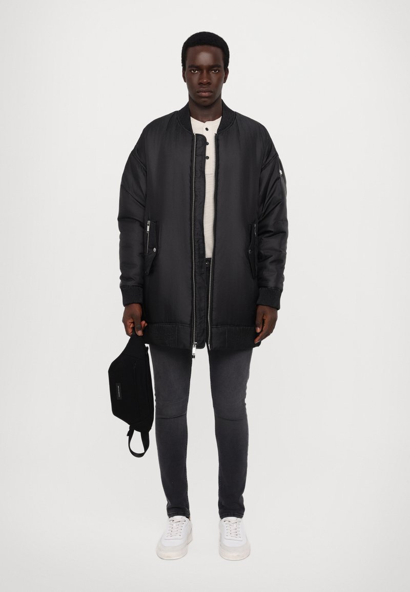 Veste bomber noire rembourrée avec des poignets en côtelé et un ourlet, portée sur une chemise henley de couleur claire. Assortie à un jean foncé et des baskets blanches.