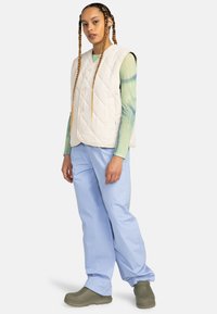 Gilet matelassé en blanc cassé avec un motif en losanges, associé à un pantalon bleu clair et des sabots vert olive. Coiffure avec de longues tresses visible.