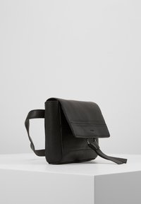 Černá kožená kabelka přes rameno s klopou a dekoračním detailem na popruhu, odložená na bílé ploše na pozadí v jednobarevné šedé barvě.