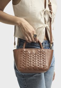 Bruine leren handtas met een geweven patroon, bovenhandvatten en een afneembare schouderriem. Logo op de voorkant weergegeven. Casual outfit met denim jeans.