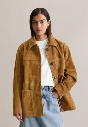 Braune Wildlederjacke mit Kragen, vorderen Knöpfen und großer Tasche. Hat eine weiche Textur und eine lässige Passform, kombiniert mit einem weißen Oberteil.