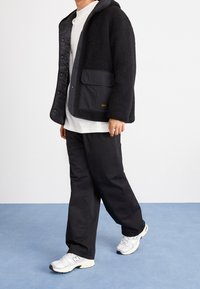 Schwarze Fleece-Jacke mit grauer Kapuze und Tasche, kombiniert mit schwarzen weit geschnittenen Hosen und weißen Turnschuhen vor blauem Hintergrund.