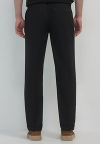Schwarze Hose mit geradem Schnitt, ausgestattet mit zwei Gesäßtaschen, umgeschlagenen Säumen und einer glatten Stoffoberfläche. Braune Schuhe sind am unteren Rand sichtbar.