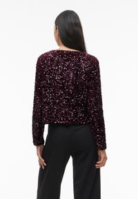 Vyno raudonio spalvos blizgučių švarkas su ilgomis rankovėmis, trumpintas kirpimas, tekstūruota paviršiaus struktūra ir apvaliu iškirptu kaklu. Apačioje matomos juodos kelnės.