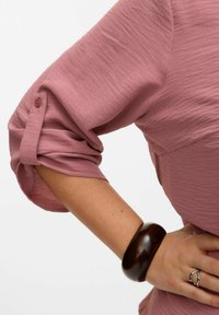 Blusa rosa con texture e maniche arrotolate, con dettagli in bottone. Un bracciale di legno scuro e un anello d'argento adornano il polso.
