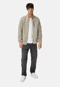 Hellbraune Zip-Jacke aus strukturiertem Stoff, mit schwarzem Reißverschluss, kombiniert mit einem weißen T-Shirt und dunkelgrauen Jeans, getragen mit weißen Sneakern.