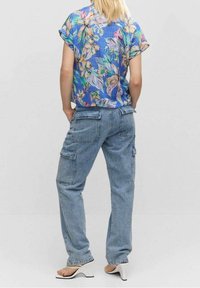 Donna con capelli lunghi biondi che indossa una camicia a maniche corte blu con motivo floreale, pantaloni cargo leggeri blu chiaro e sandali con tacco bianchi, in piedi con le mani nelle tasche.