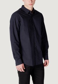 Camicia blu navy con piccoli pois bianchi, maniche lunghe e colletto classico. Il tessuto sembra liscio e leggero.