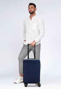 Maleta rígida de color marino con textura suave, mango telescópico y cuatro ruedas multidireccionales, situada al lado de un hombre con ropa ligera.