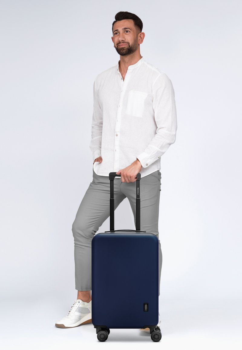 Maleta rígida de color marino con textura suave, mango telescópico y cuatro ruedas multidireccionales, situada al lado de un hombre con ropa ligera.