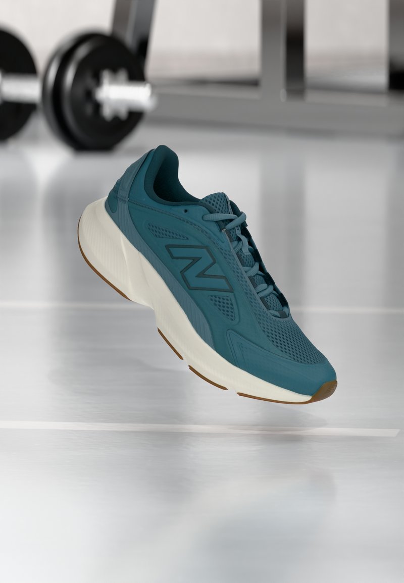 Τσάι-πράσινο αθλητικό παπούτσι με λευκή σόλα που αιωρείται εσωτερικά, βάρη γυμναστηρίου και αντανακλαστικό δάπεδο σε απαλό θολό φόντο.