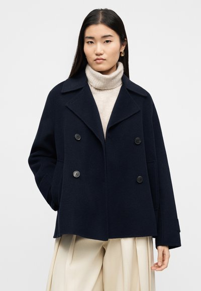 Γυναίκα που φοράει ένα διπλόμπουτο ναυτικό παλτό πάνω από ένα μπεζ πουλόβερ ζιβάγκο και μπεζ παντελόνι με ευρύ πόδι, στέκεται μπροστά σε απλό φόντο.