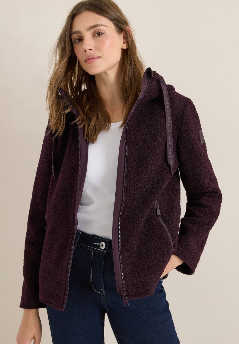 Veste en polaire bordeaux avec fermeture éclair, deux poches latérales et capuche. Présente une doublure lisse et un détail à cordon, portée par-dessus une chemise blanche.