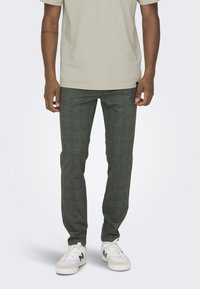 Donkergroene geruite broek van lichtgewicht stof, met een getailleerde pasvorm, gecombineerd met witte sneakers en een beige t-shirt.