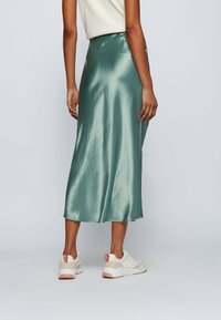 Keskipitkä, kiiltävä satiinihame vaaleanvihreänä, jossa on sileä rakenne ja laaja siluetti, yhdistettynä kermaisiin lenkkareihin.