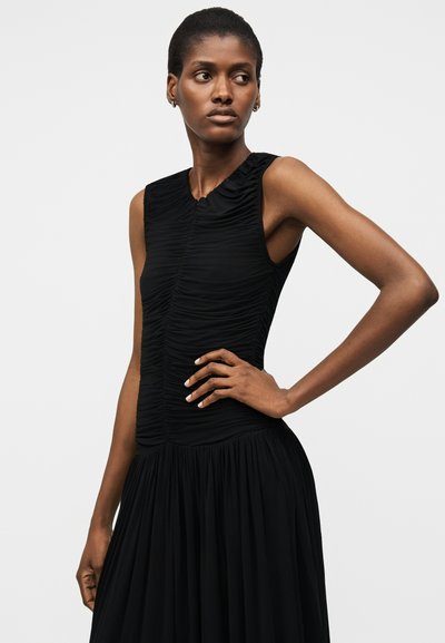 Lühikeste juustega naine, kes kannab äravoltimata musta kleiti, käsi puusal, vaadates kõrvale helel taustal.