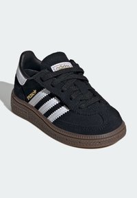 Black suede sneaker with white stripes, brown rubber sole, and black laces. Features textured upper and gold branding on tongue.