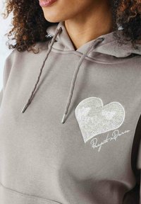 Sudadera gris con un diseño bordado en forma de corazón en un beis claro, con ojales y cordones metálicos. Textura suave.