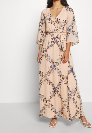 Femme portant une robe portefeuille beige à motifs floraux avec manches cloche trois-quarts, une ceinture à la taille et des sandales blanches à talons ouverts, debout devant un fond blanc.