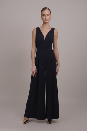 Marineblauer Jumpsuit mit V-Ausschnitt, ärmellosem Design, weiten Beinen, Falten an der Taille und glatter Textur. Kombiniert mit goldenen spitz zulaufenden Schuhen.