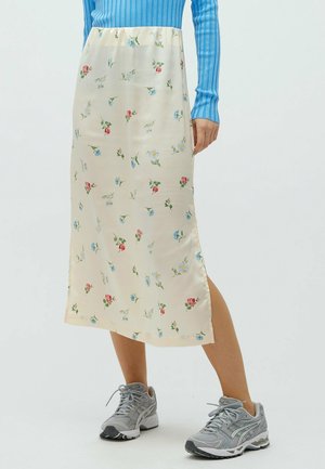 Midilanger cremefarbener Rock mit kleinen roten, blauen und gelben Blumen, kombiniert mit grauen Sneakers und einem blauen, gerippten Langarmshirt.