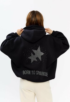 Schwarzer oversized Hoodie mit Kapuze, verziert mit einem Stern aus Strasssteinen und dem Schriftzug "BORN TO SPARKLE" auf der Rückseite. Weiche Stofftextur.