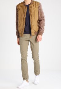 Brązowa kurtka typu bomber z skórzanymi rękawami, granatowy t-shirt, khaki spodnie i białe trampki. Kurtka posiada kieszeń na zamek na rękawie.