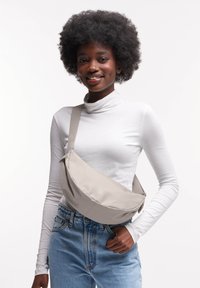 Borsa a tracolla in tessuto grigio chiaro con forma curva, tracolla regolabile e dettaglio del logo, indossata sopra un dolcevita bianco e jeans blu.