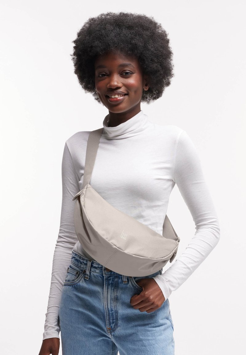 Borsa a tracolla in tessuto grigio chiaro con forma curva, tracolla regolabile e dettaglio del logo, indossata sopra un dolcevita bianco e jeans blu.