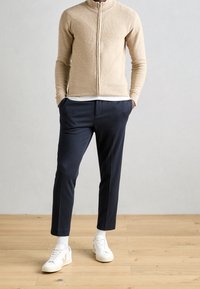 Beżowy, zapinany na zamek sweter z dzianiny z długim rękawem, zestawiony z dopasowanymi, ciemnogranatowymi spodniami i białymi trampkami. Tło z drewnianą podłogą.