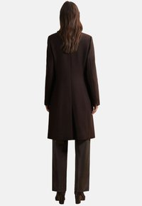 Abrigo de lana marrón con corte recto, mangas largas y un dobladillo de longitud media. Combinado con pantalones oscuros y botas de tacón.