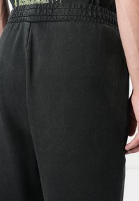 Svart joggebukse laget av myk, teksturert stoff med elastisk midjebånd og sidelommer, vist fra baksiden, med avslappet passform.