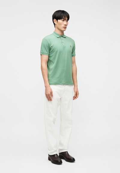 Άνδρας που φοράει πουκάμισο πόλο σε χρώμα φασκόμηλο, λευκό παντελόνι και σκούρα καφέ  μπότες, στέκεται μπροστά σε απλό λευκό φόντο.