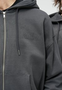 Hanorac gri cu fermoar, realizat din țesătură moale. Dispune de glugă cu șnur, buzunare frontale, marcă în relief pe piept și mâneci lungi.