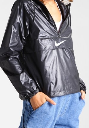 Persona que lleva una cazadora cortavientos negra de Nike con media cremallera y vaqueros azules, manos en los bolsillos, mostrada desde los hombros hasta las caderas.